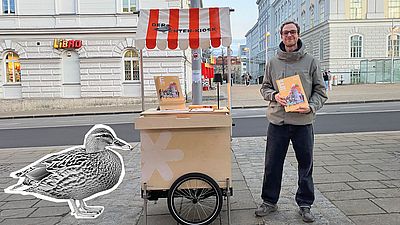 Ein Mann steht auf einem städtischen Gehweg neben dem kleinen mobilen ENTE Mitmach-Magazin Kiosk mit rot-weiß gestreiftem Dach und hält ein Magazin in den Händen. Auf dem Kiosk liegen weitere Exemplare aus, während im Hintergrund helle, klassische Gebäude eine ruhige Straßenszene bilden. Links im Bild ist ein ausgeschniettenese schwarzweisfoto einer Ente.