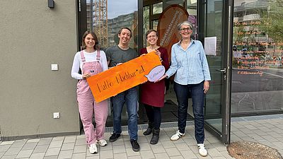 Vier GB*Mitarbeiter*innen vor dem GB*Stadtteilmanagement halten herzlich lachend ein Schild mit der Aufschrift "Willkommen!" in die Kamera.