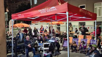 Personen sitzen auf Stühlen unter einem Pavilion auf der Straße und schauen auf eine Leinwand.
