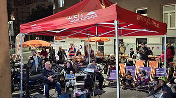 Personen sitzen auf Stühlen unter einem Pavilion auf der Straße und schauen auf eine Leinwand.