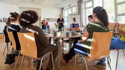 GB*Frauencafé 8 Frauen sitzen auf Sesseln rund um einen Tisch und sehen aufmerksam einer Vortragenden zu, die beim Flipchart steht