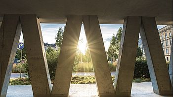 Sonnenuntergang, der zwischen schräg zulaufenden Betonpfeilern durchstrahlt. Im Hintergrund ist die Innenhofbegrünung sichtbar.