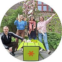 Vier GB*Mitarbeiter*innen rund um eine Schatzkiste werfen lachend übergroße Mützen in die Luft