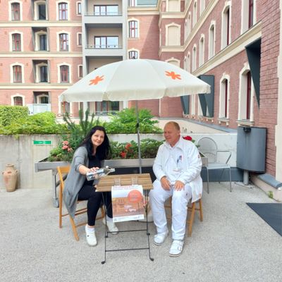Oberarzt und Frau auf Klappsesseln an einem Klapptisch sitzend unter einem gebrandeten Sonnenschirm sitzend. Vom Tisch hängt ein laminiertes Plakat runter, das darauf hinweist, dass dieses Setting ein Sommer-Rastplatz ist.