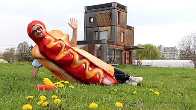 Eine Person, verkleidet als Hotdog lädt freundlich zum Nachbarschaftstreffen.