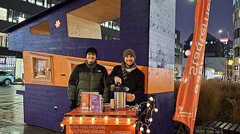 Zwei Personen stehen beim GB*Infopoint und schenken Kaffee aus.