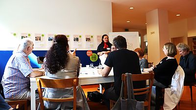 ca. 6 Personen sitzen im Innenraum an Tisch und sprechen