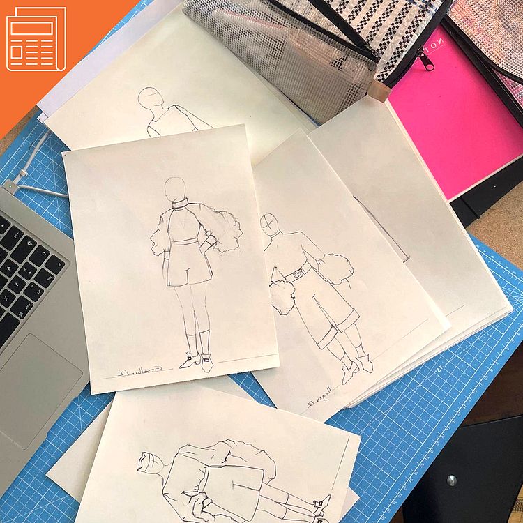 Ein aufgeklappter Laptop und Skizzen von Kostümen liegen auf einem Tisch.