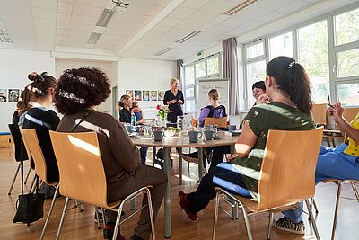 8 Frauen sitzen auf Sesseln rund um einen Tisch und sehen aufmerksam einer Vortragenden zu, die beim Flipchart steht
