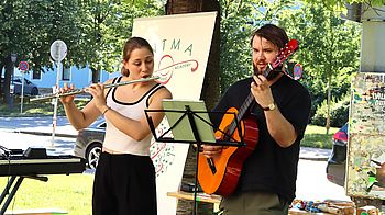 zwei Personen stehen unter Baum, eine Person spielt Querflöte, eine Person Gitarre. Sommerliche Stimmung