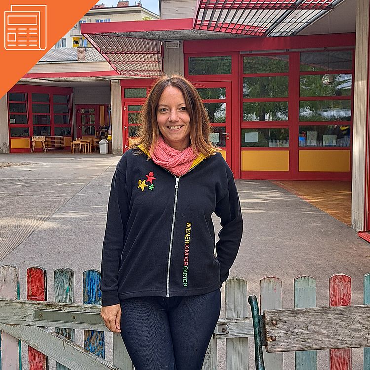 Das Bild zeigt eine Frau, die vor einem Kindergarten mit roten und gelben Elementen steht. Sie trägt eine schwarze Jacke mit der Aufschrift "Wiener Kindergärten" in bunten Buchstaben und hat einen rosa Schal um den Hals. Im Hintergrund sieht man einen offenen Eingangsbereich mit großen Glasfenstern, durch die Tische und Stühle zu erkennen sind. Der Außenbereich ist von einem kleinen Holzzaun umgeben, der teilweise bunt bemalt ist. Die Frau lächelt in die Kamera.