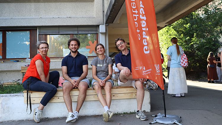 vier Personen, zwei männlich zwei weiblich gelesen, weiß, sitzen auf Bank. orangener Banner