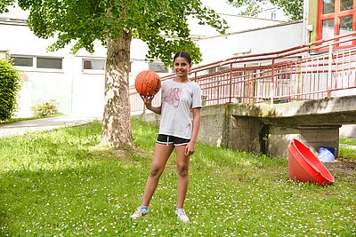 Angela steht in einer Wiese und hält einen Basketball in der Hand.