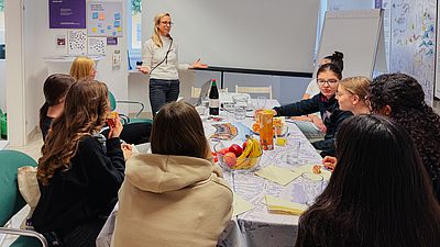 Mädchen an Tisch vor Mitarbeiterin der GB*, die über Stadtplanung referiert