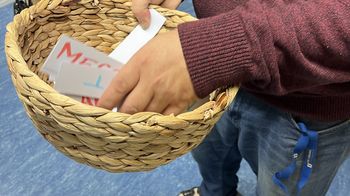 Eine Person hält einen kleinen Korb in ihrer rechten Hand. Mit der linke zieht sie einen Zettel, worauf ein Begriff steht.