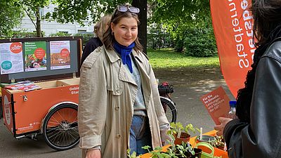 Frau hinter orangenem Tisch, darauf viele Setzlinge.
