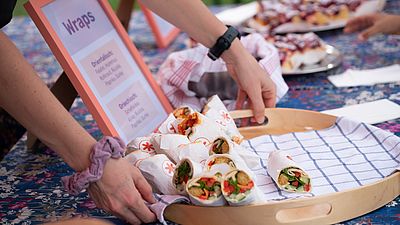 Zwei Hände halten ein Tablett auf dem mehrere Wraps angeordnet sind.