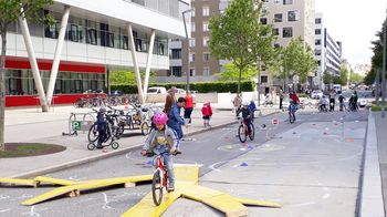 In einer Fußgängerzone ist ein Radparcours aus gelben Holzplanken aufgebaut, KInder fahren mit Fahrrädern darüber