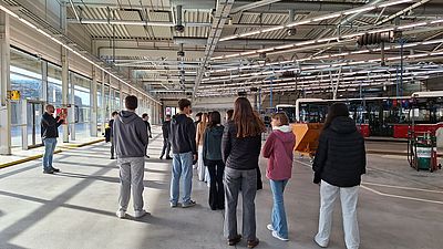 mehrere junge Personen stehen in großer Halle mit Bussen