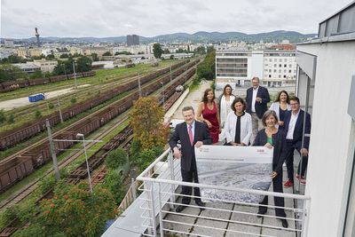 8 PolitikerInnen stehen auf einem Balkon und halten einen Plan, im Hintergrund Bahnschienen