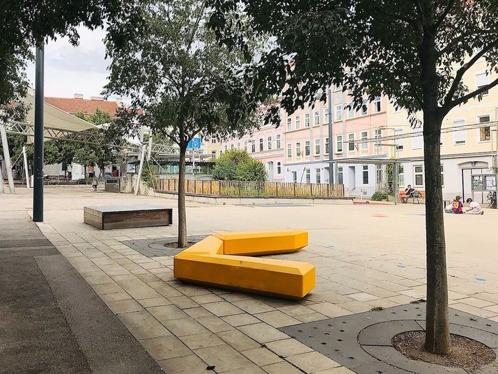 Öffentlicher Platz mit Bäumen und einem gelben Sitzelement in der Mitte