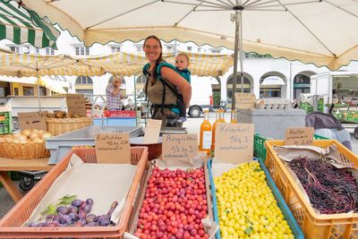 Carmen Leiner vor ihrem Stand, buntes Obst, Leiner trägt Kind am Rücken