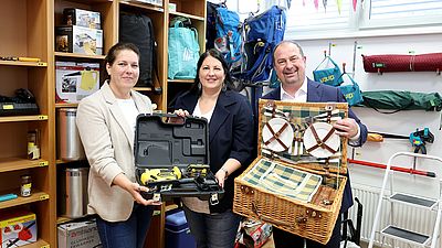 StRIn Kathrin Gaál, BVIn Christine Dubravac-Widholm und BV Alexander Nikolai halten eine Bohrmaschine und einen Picknickkorb aus dem neuen Leihladen hoch.