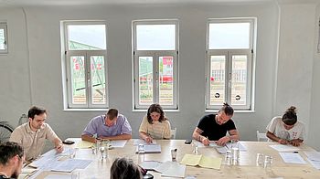 Teilnehmende sitzen um großen Tisch im Stadtraum und schreiben.