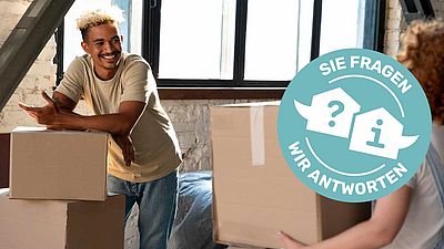 Junger Mann stütz sich auf Umzugskartons ab und strahlt junge Frau, die Umzugskarton trägt, an.
