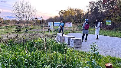 Grüne Fläche mit Pflanzne, Infoschildern, drei Personen stehend