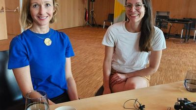 Zwei Frauen sitzen und lächeln in die Kamera.