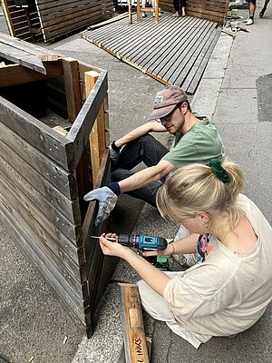 Ein Mann hält ein Brett und eine Frau schraubt eine Schraube mit einem Akkuschrauber hinein.
