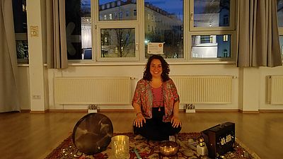Eine Frau sitzt auf einem Tepich umgeben von Klangschalen, Duftölen und Lichterketten.
