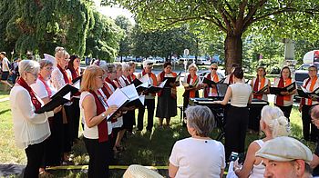Chor mit ca. 25 Personen, meist weiblich gelesen und weiß, steht im Park mit Notenbüchern. Publikum sitzend 3 Personen