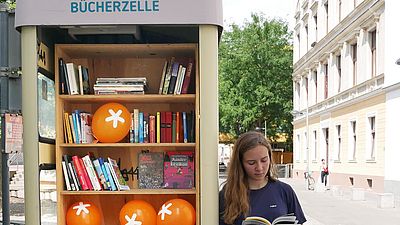 Telefonzelle gefüllt mit Büchern. Rechts steht eine Frau, die in einem Buch blättert.