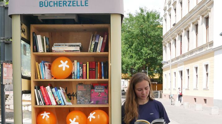 Telefonzelle gefüllt mit Büchern. Rechts steht eine Frau, die in einem Buch blättert.
