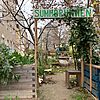 Das Foto zeigt einen Gemeinschaftsgarten mit dem Namen "Summbrunnen", wie auf dem handbemalten Schild am Eingang zu lesen ist. Der Eingang ist durch einen Holzrahmen mit grüner Beschriftung markiert. Links und rechts des Eingangs befinden sich Hochbeete aus Holz, die mit verschiedenen Pflanzen und Kräutern bepflanzt sind. Ein kleiner Weg führt tiefer in den Garten, der von weiteren Beeten, Bäumen und Sträuchern geprägt ist.  Im Hintergrund sind Sitzgelegenheiten und Gartenmöbel erkennbar, die einen einladenden Eindruck vermitteln. Die Szenerie wirkt naturbelassen und gemütlich, mit sichtbaren Elementen von Nachhaltigkeit, wie der Einsatz von Holzpaletten und Upcycling-Materialien. Im Hintergrund sieht man ein städtisches Umfeld mit Gebäuden und kahlen Bäumen, was auf eine Herbst- oder Winterzeit hinweist. Der Garten scheint ein Ort der Begegnung und des urbanen Grüns zu sein.