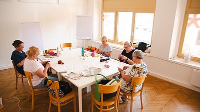 5 Personen sitzen an einem Tisch, stricken und plaudern.