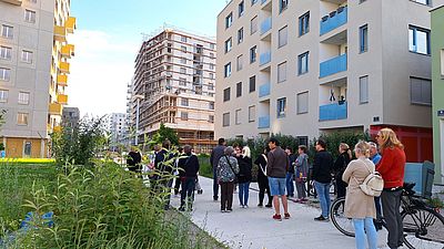 ca. 20 Personen gehen zwischen hohen grauen Gebäuden