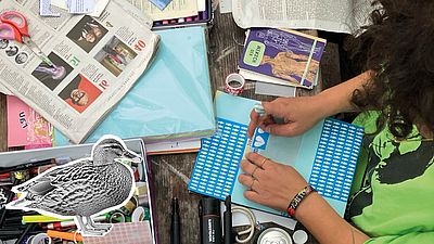 Person gestaltet an einem Tisch mit Papier, Stiften und ausgeschnittenen Bildern eine Collage; im Vordergrund liegt eine schwarz-weiße Entenillustration.