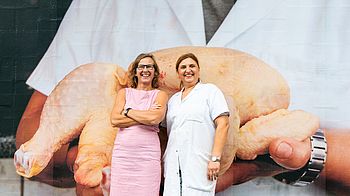 Leben am Schlingermarkt, Plakate am Markt, Leiterin der GB* und Standlerin vor Plakat mit Hühnchen