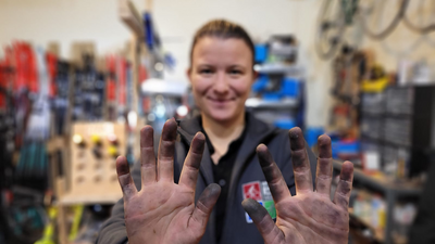 Eine Frau hält ihre schmutzigen Hände hoch, im Hintergrund sieht man eine Werkstatt.