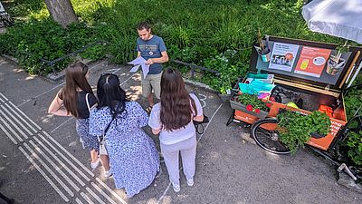 3 Jugendliche machen bei einem Quiz mit. Vor ihnen steht ein GB*Mitarbeiter und stellt ihnen die Fragen, die er auch auf einem Blatt Papaier ausgedruckt hat. Rechts neben ihnen steht das mit Blumenkisten geschmückte GB*Lastenrad