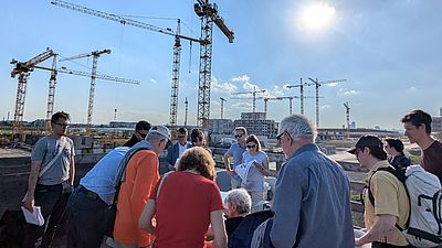 Mehrere Menschen stehen outdoor um einen Tisch mit Planungsunterlagen herum, im Hintergrund sind Baukräne zu sehen.