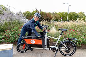 Person steht in Park mit Lastenfahrrad und holt etwas vorne aus der Ladefläche