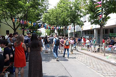 Das Bild zeigt ein lebendiges Straßenfest mit vielen Menschen. Kinder und Erwachsene basteln an Tischen, bunte Papierlaternen hängen über der Straße. Die Stimmung ist fröhlich und einladend.