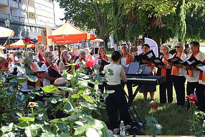 Chor mit ca. 25 Personen, meist weiblich gelesen und weiß, steht im Park mit Notenbüchern. Rosen im Vordergrund