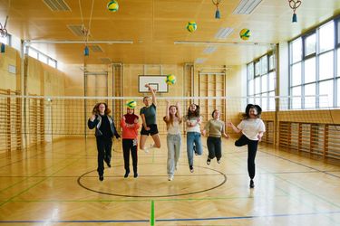 7 Kinder springen in die Luft in einem großen Turnsaal und werfen Volleybälle in die Luft.