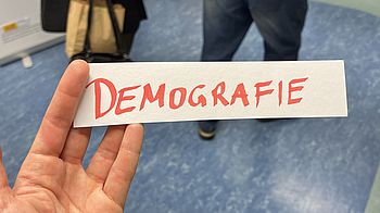 Eine rechte Hand hält einen Zettel mit der Aufschrift "Demografie" in Großbuchstaben.