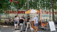 Um einen Stehtisch mit Orange-weißen Schirm stehen mehrere Personen auf einem öffentlichen Platz im Sommer